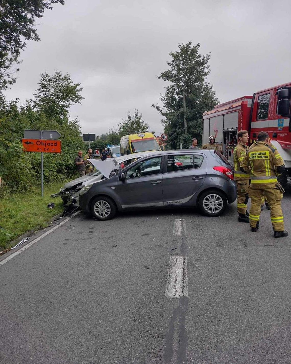 Na zdjęciu widać dwa uszkodzone pojazdy, które brały udział w wypadku, a także karetkę pogotowia, radiowóz policyjny i wóz strażacki wraz ze strażakami.